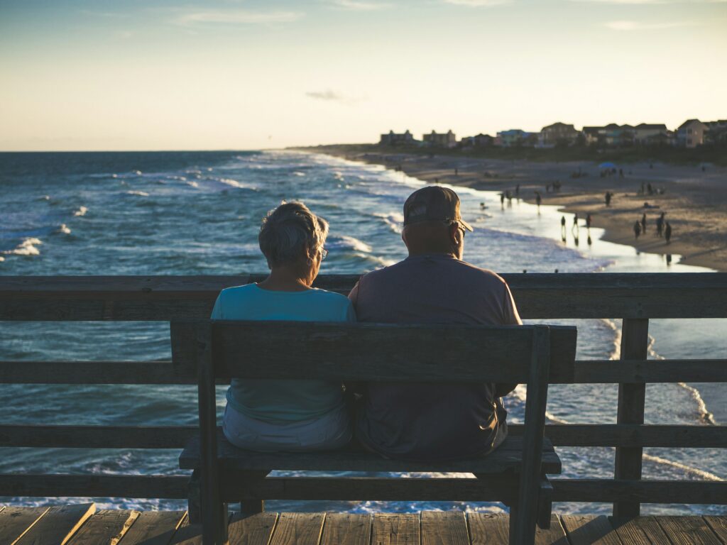 Back view of two people sitting on a wooden railing on a pier, looking at the ocean at sunset.