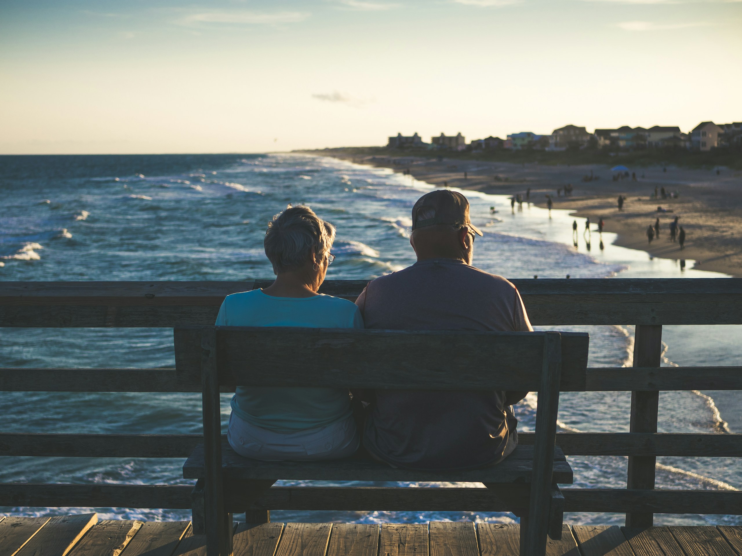 Back view of two people sitting on a wooden railing on a pier, looking at the ocean at sunset.