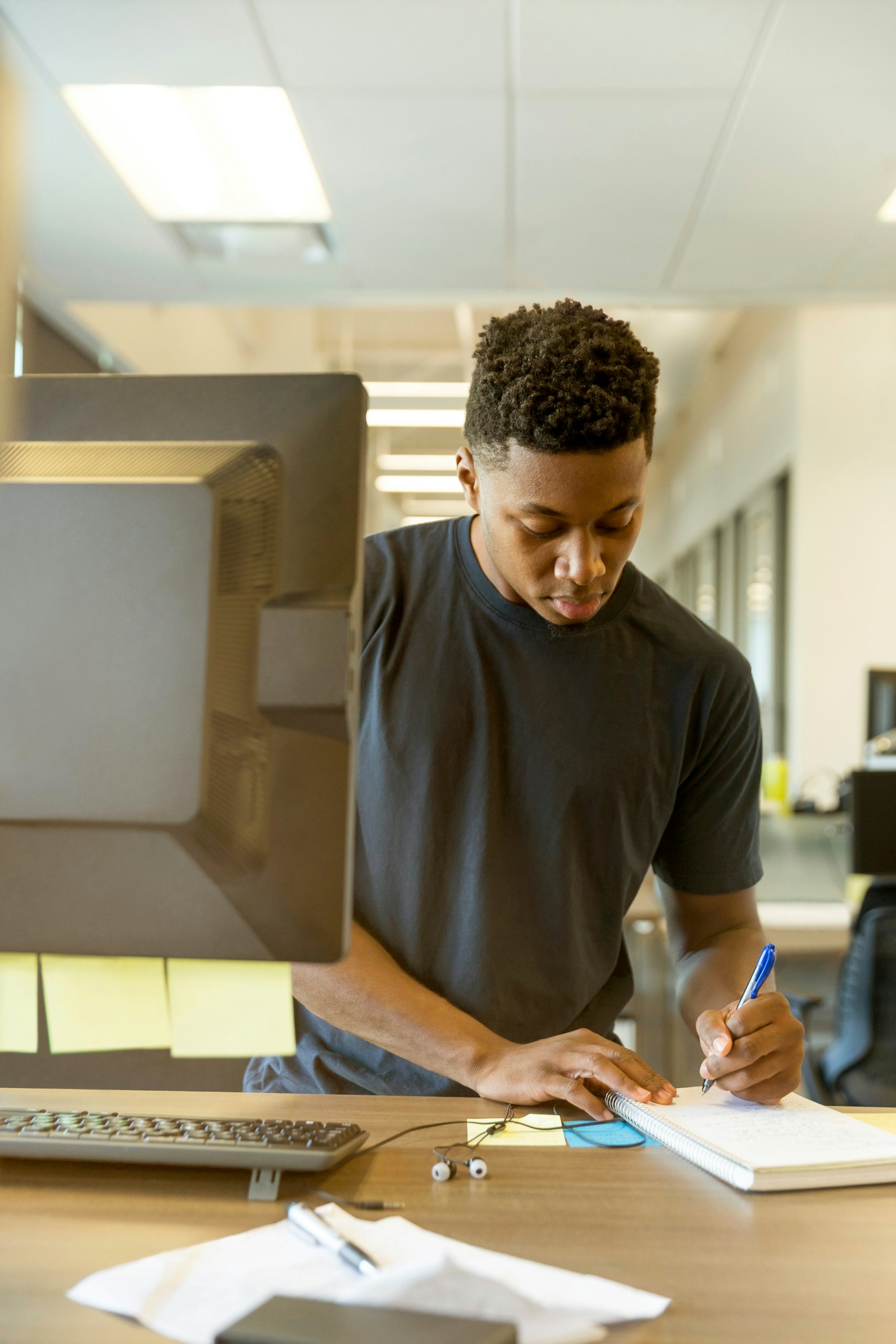 A person in a dark T-shirt writes in a notebook at a desk, with a computer monitor, keyboard, and sticky notes nearby.