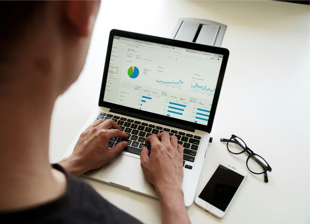 Person typing on a laptop showing analytics dashboards on screen (pie chart, charts) with a smartphone and glasses nearby