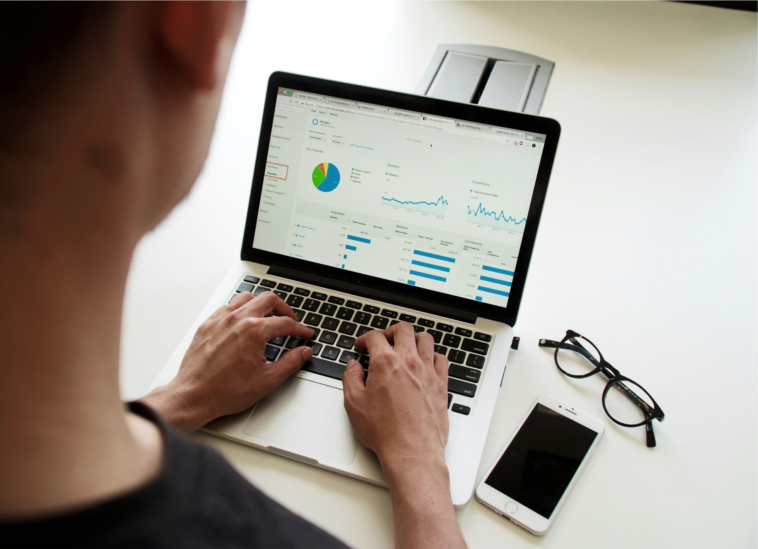 Person typing on a laptop showing analytics dashboards on screen (pie chart, charts) with a smartphone and glasses nearby