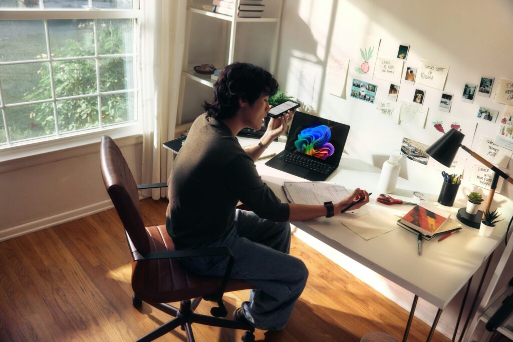 Person sitting at a white desk in a bright home office, using a smartphone while a colorful laptop screen glows nearby.
