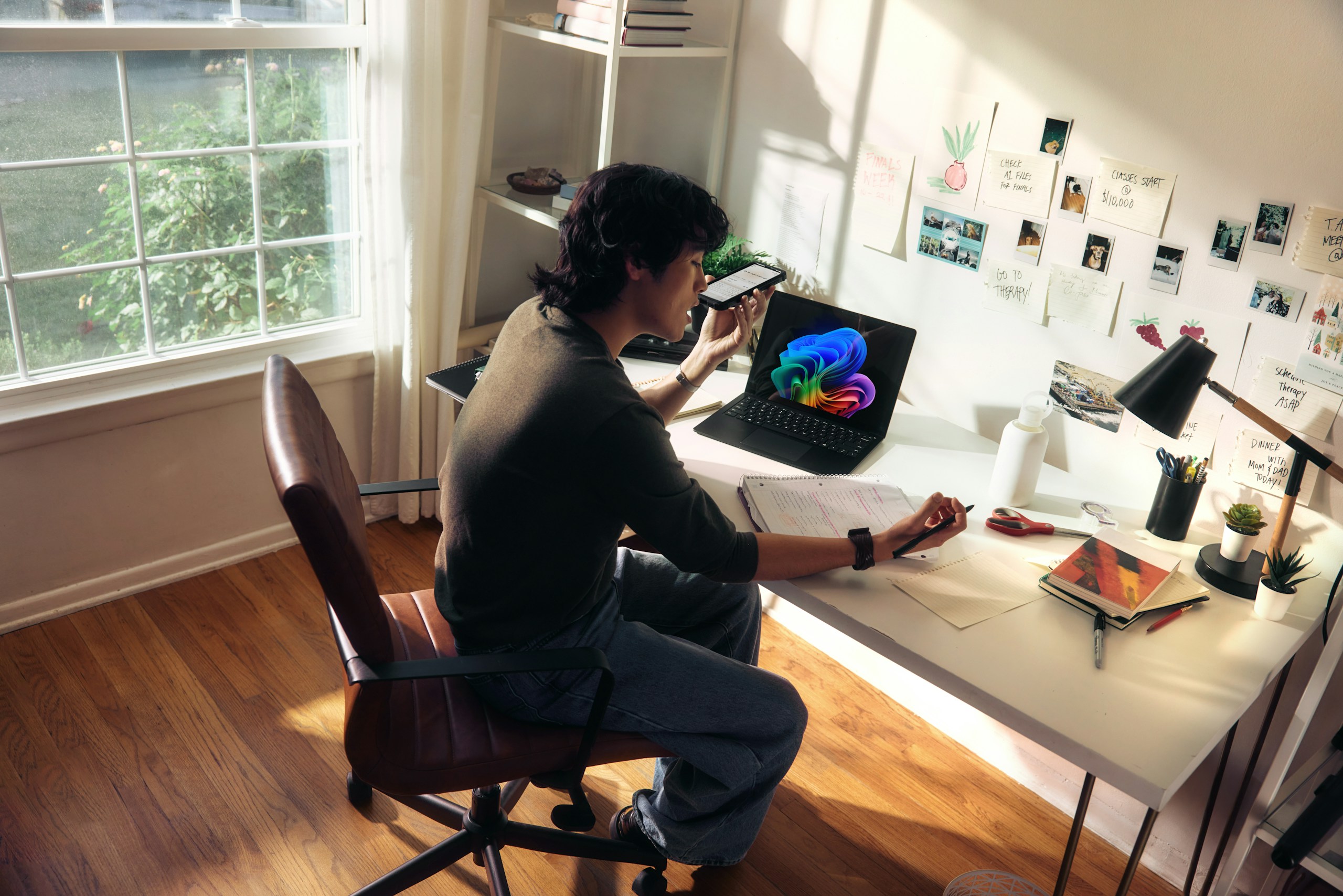 Person sitting at a white desk in a bright home office, using a smartphone while a colorful laptop screen glows nearby.