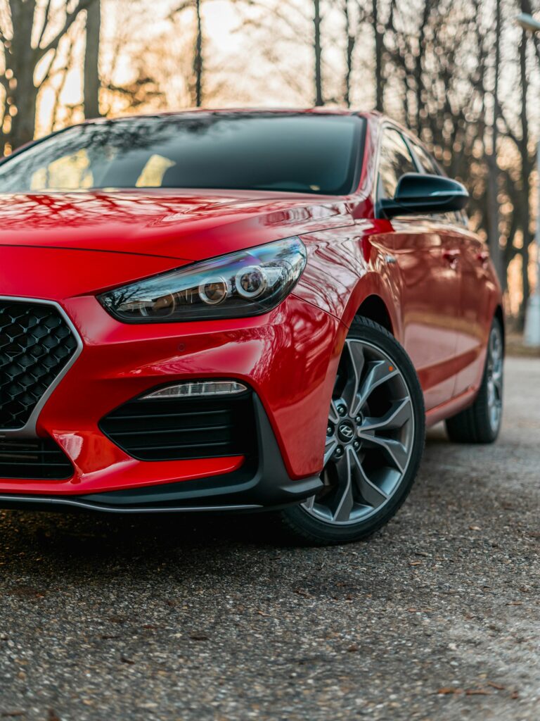 Front-left view of a red sedan, showing the headlight, grille, and glossy body with trees in the background.