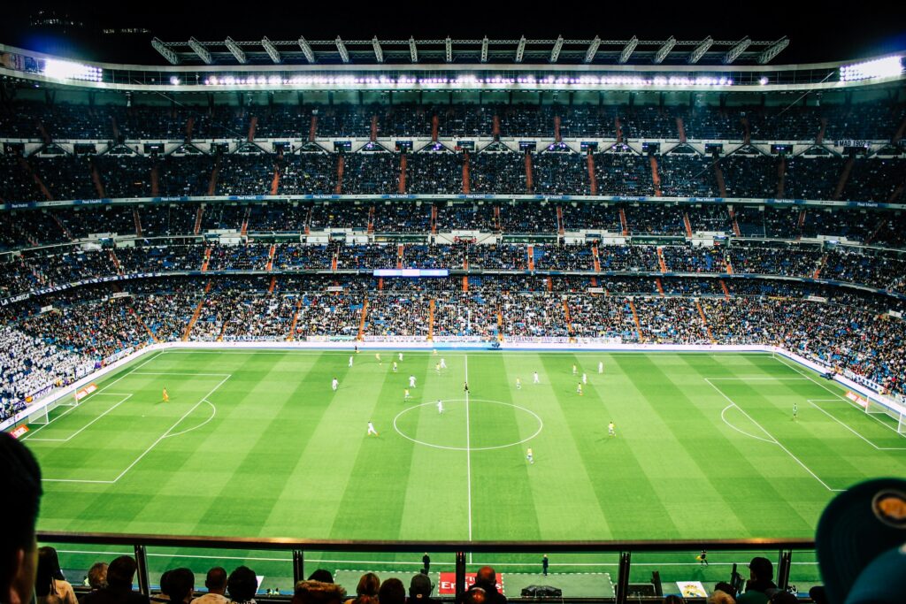 Nighttime soccer match in a large packed stadium with players on the green field and a full stands crowd amid bright floodlights in the background,
