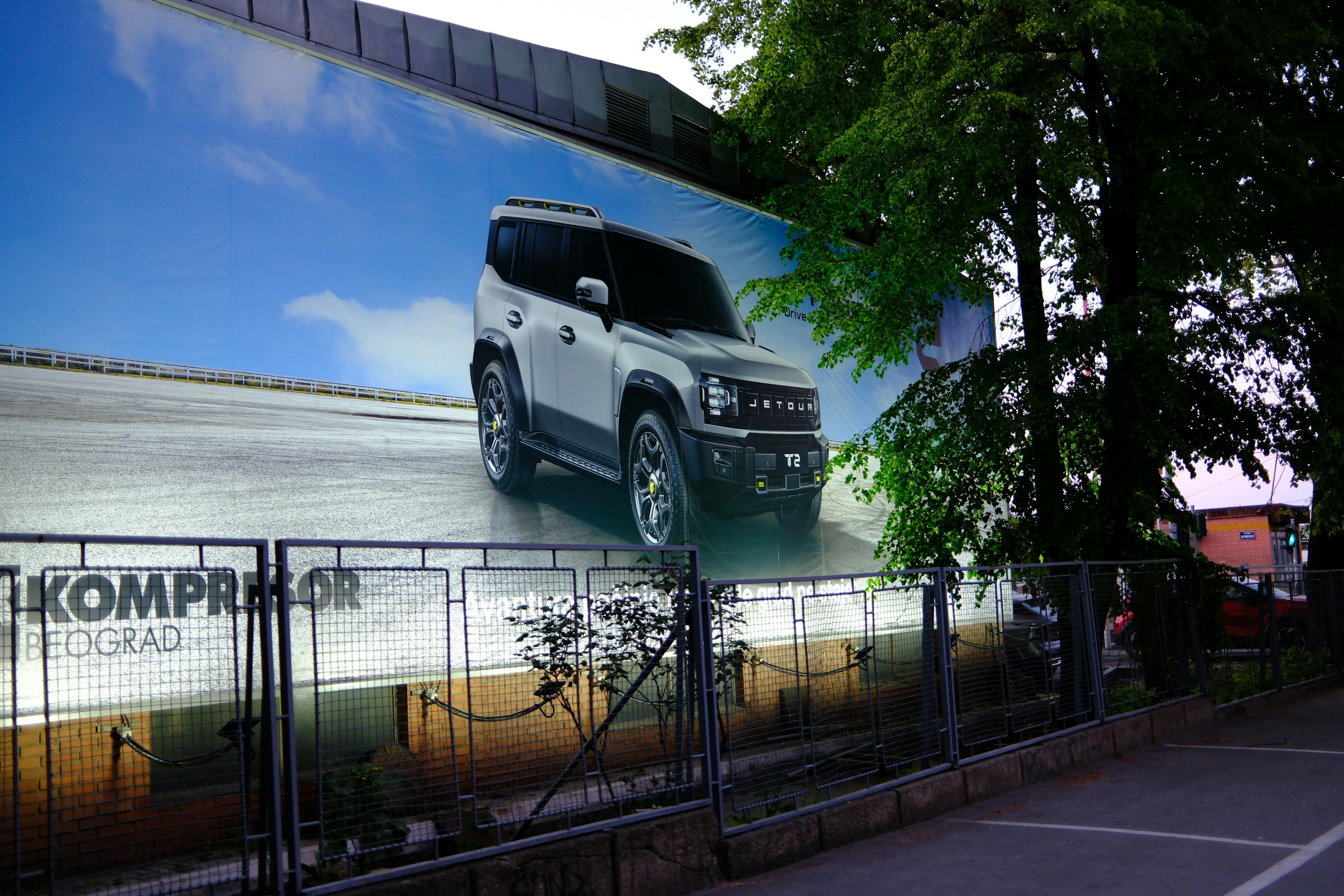 Large outdoor billboard featuring a white Jetour SUV against a blue sky, with a metal fence and tree line in the foreground.