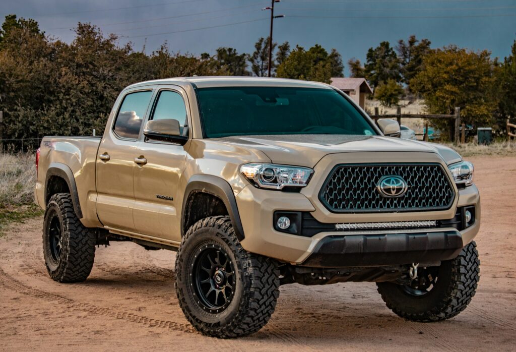 Front three-quarter view of a beige Toyota Tacoma pickup with large off-road tires on a dirt road