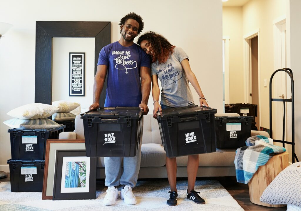 Two people stand behind large black Hive Boxx storage bins in a living room, smiling at the camera with framed art and pillows nearby, as if packing or moving belongings.