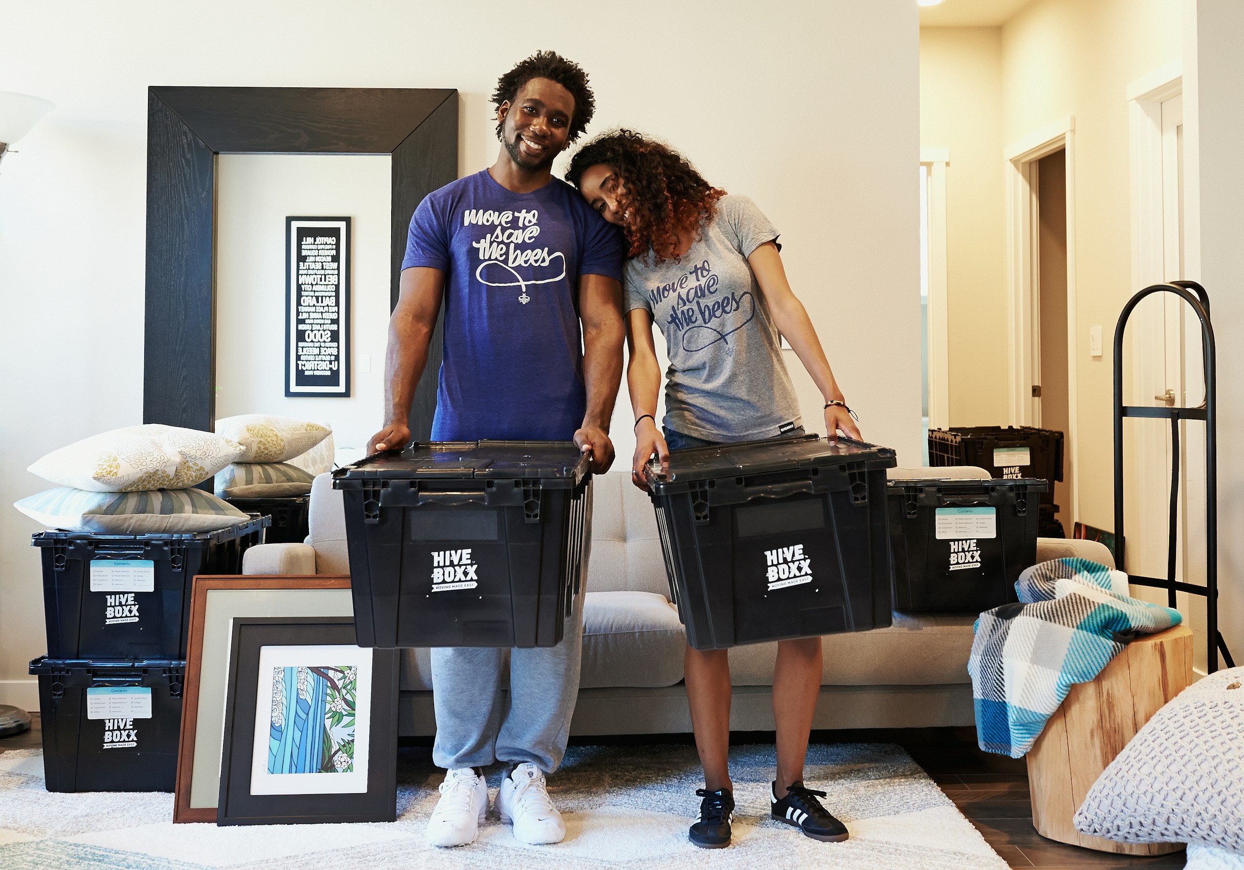 Two people stand behind large black Hive Boxx storage bins in a living room, smiling at the camera with framed art and pillows nearby, as if packing or moving belongings.