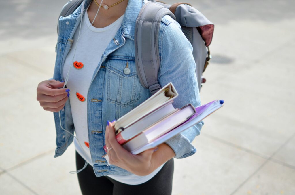 Student wearing a denim jacket over a white shirt with watermelon patches, carrying notebooks and a backpack with earbuds.