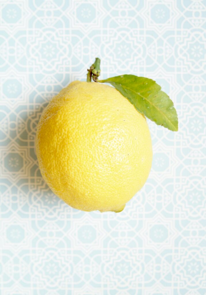 Close-up of a bright yellow lemon with a green leaf against a pale blue geometric patterned background, capturing texture and color.
