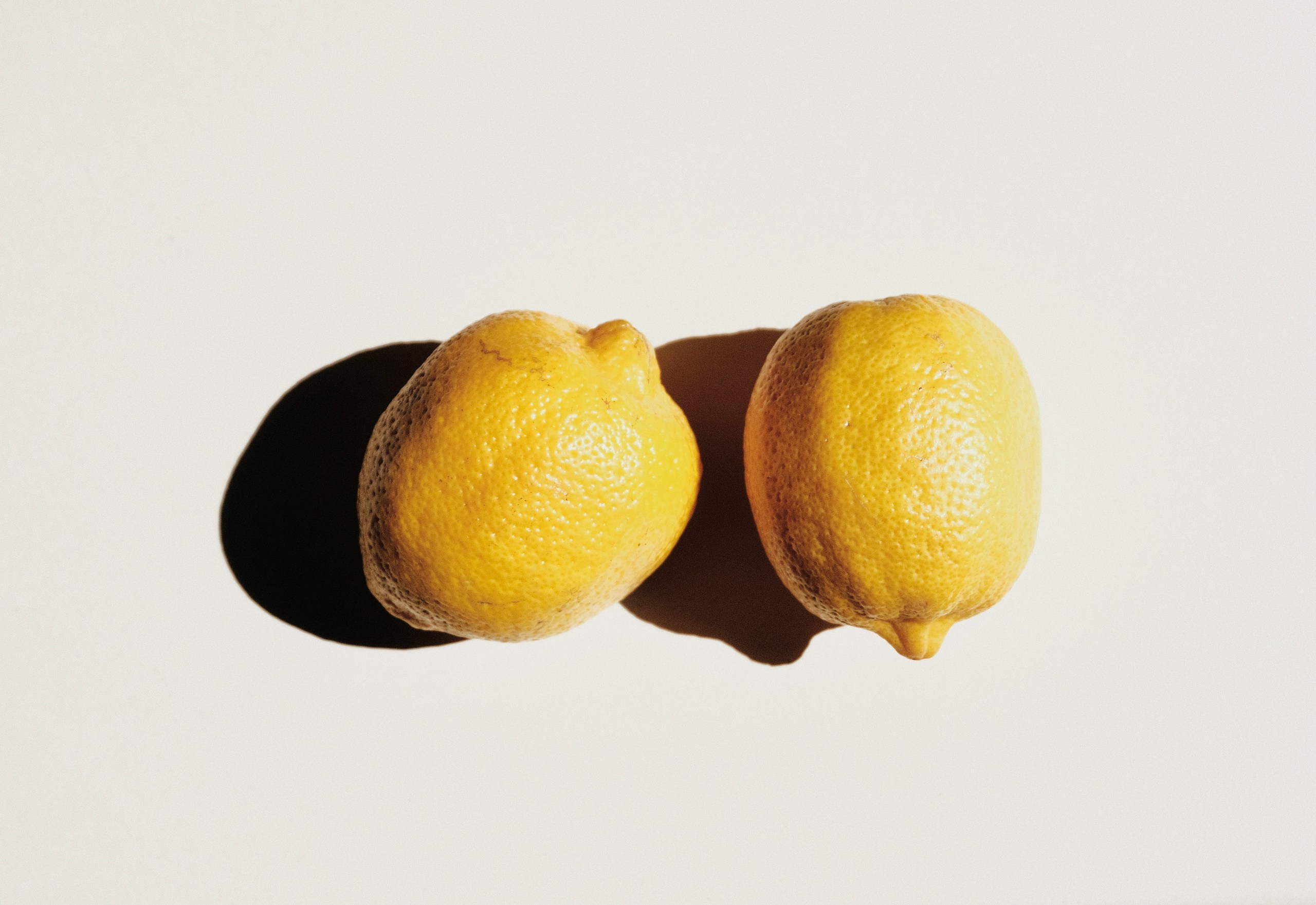 Two bright yellow lemons resting together on a pale background with a soft shadow nearby