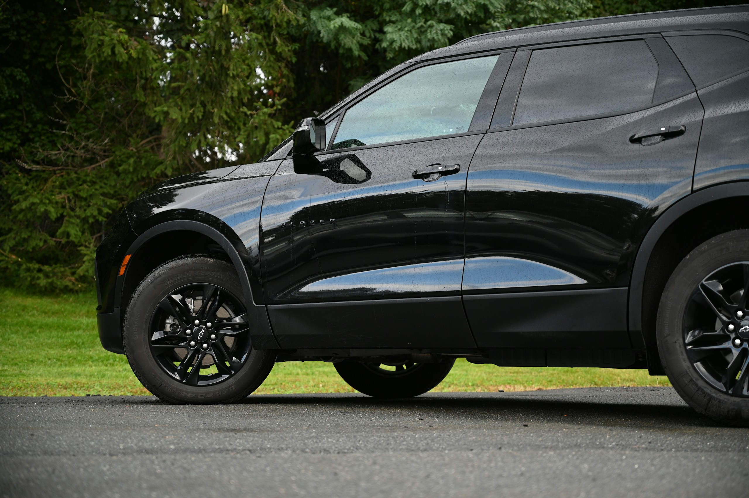 Close-up side view of a black SUV with raindrops on its surface, parked on a paved road with green trees in the background.
