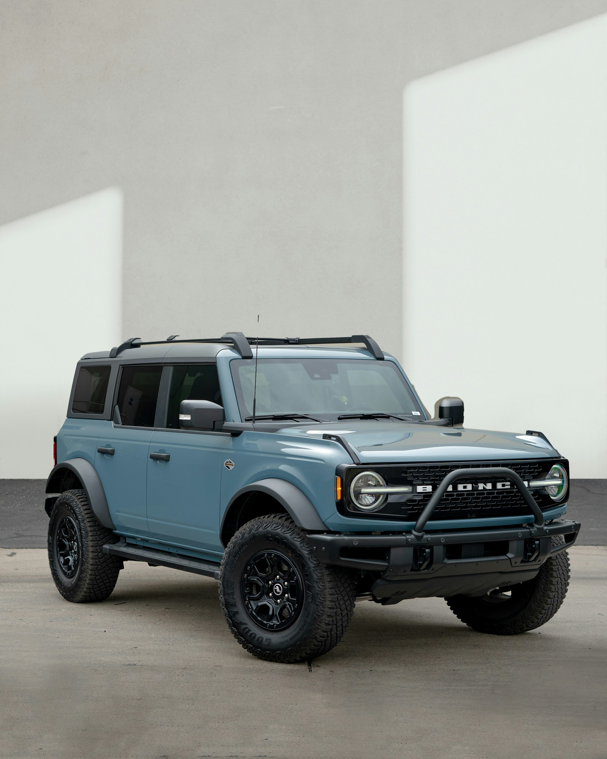 Front three-quarter view of a blue Ford Bronco with black off-road tires and roof rack in a parking lot.