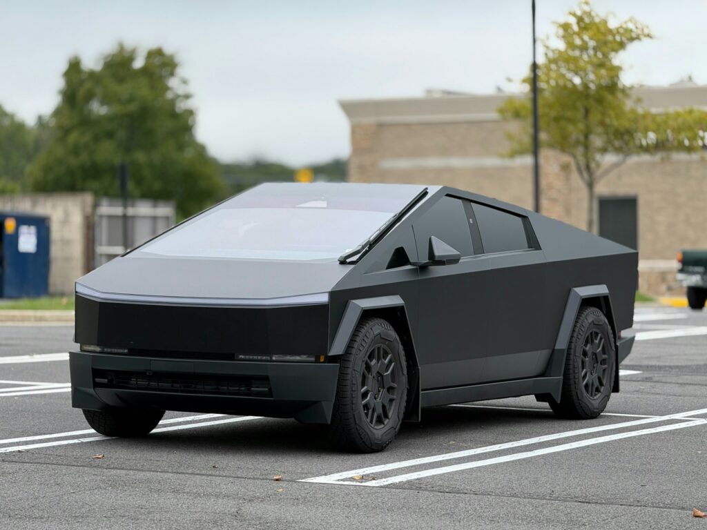 Futuristic matte black armored-looking car with angular panels parked in a striped lot.