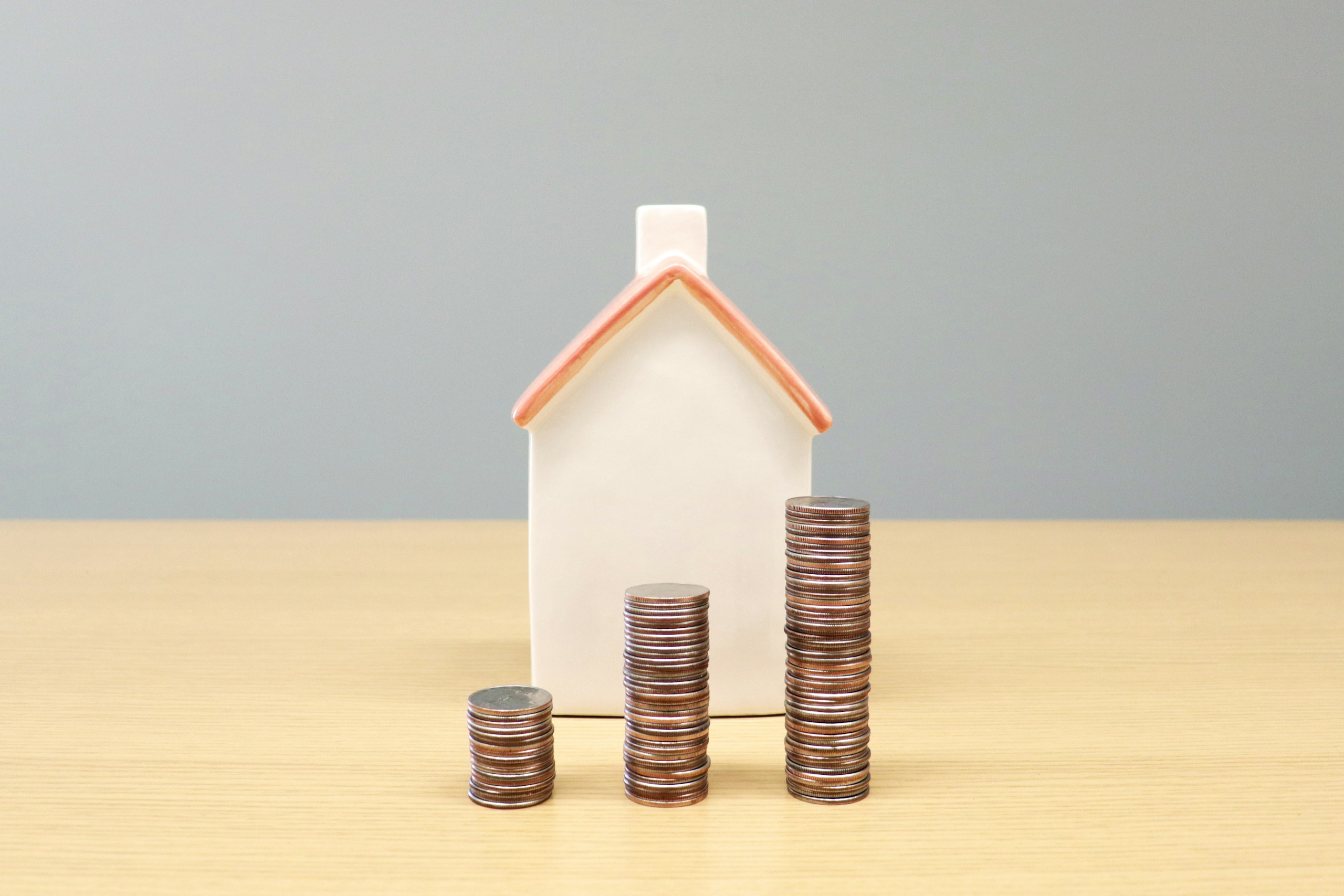 White house model behind three stacks of coins of increasing height on a wooden table, illustrating savings or home finance growth.