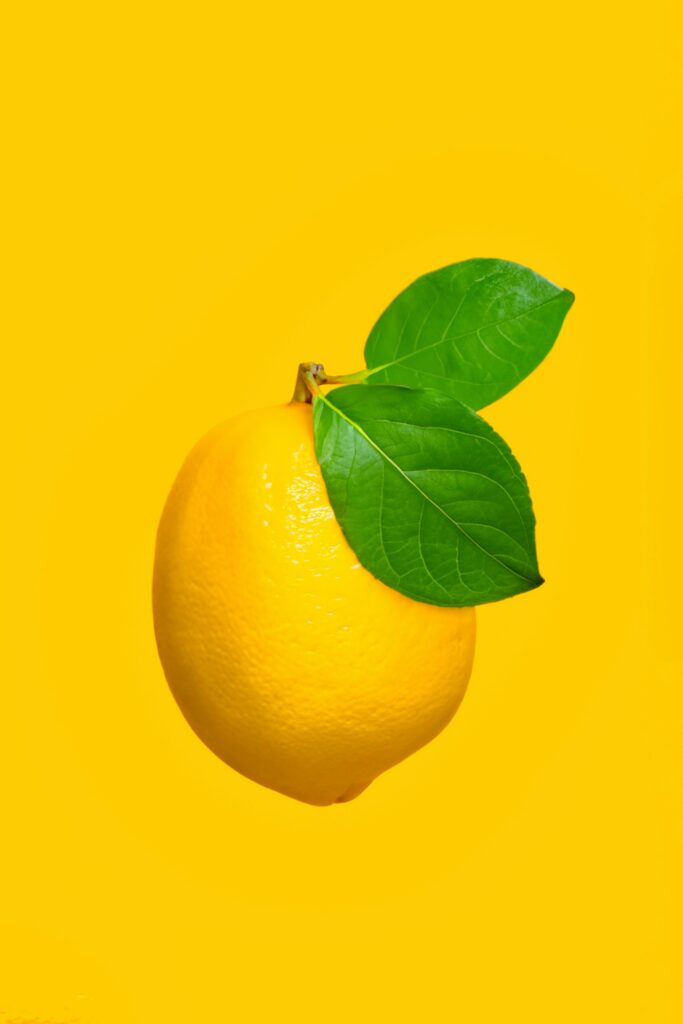 Yellow lemon with green leaves against a solid bright yellow background.