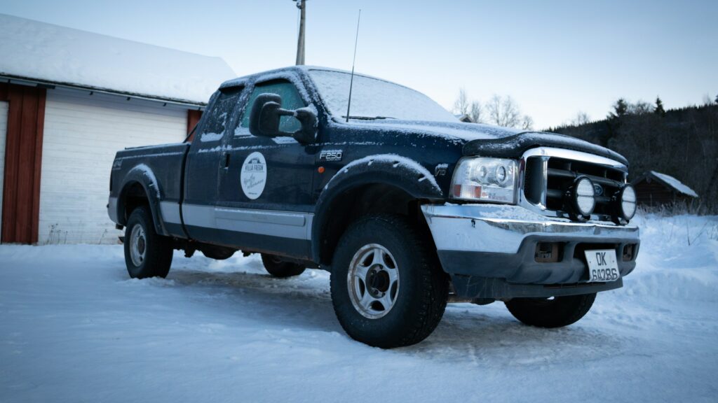 Blue Ford F-250 pickup truck covered in snow parked on a snowy driveway with a white building in the background.