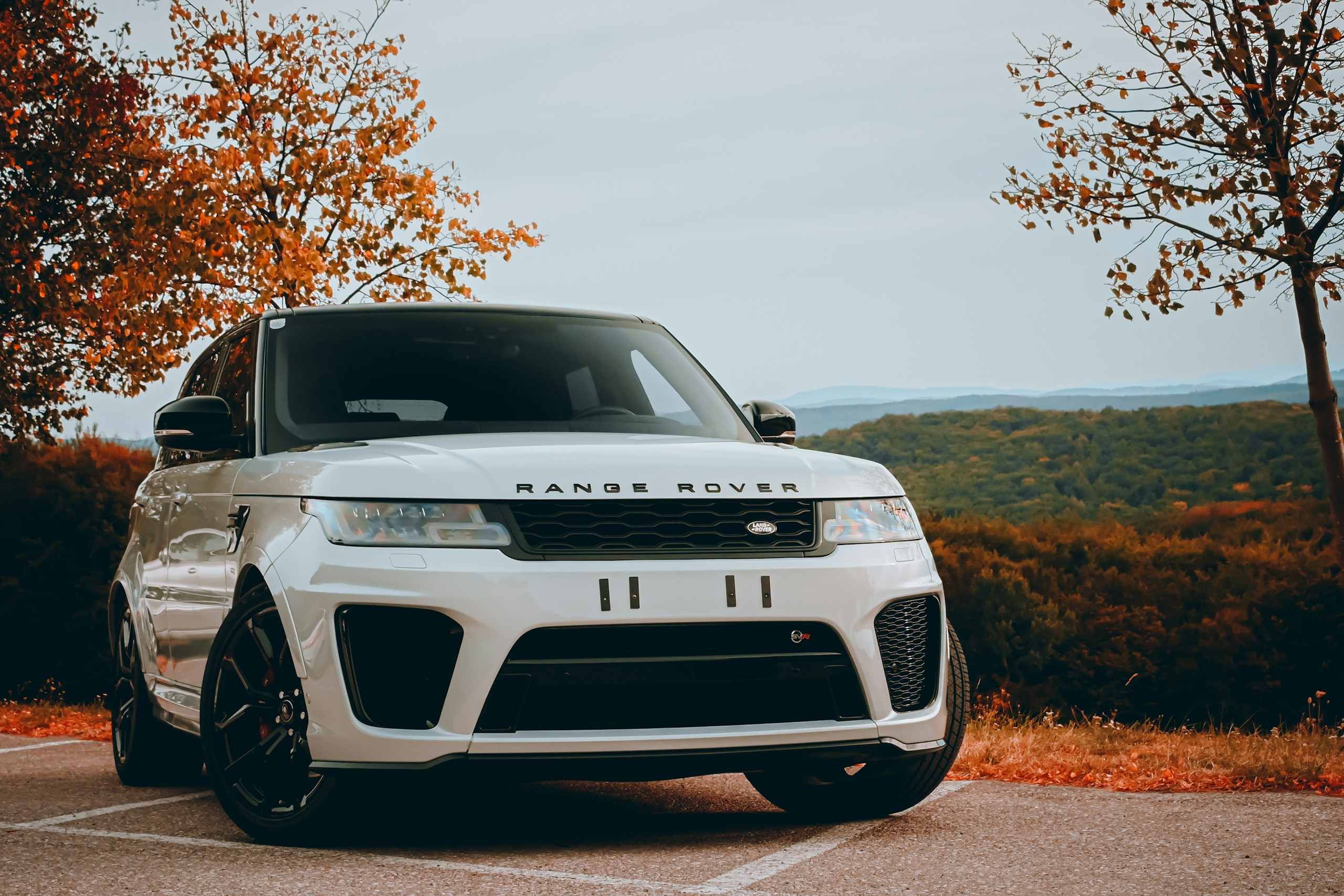 White Range Rover SUV parked on a road with autumn trees and distant hills in the background.