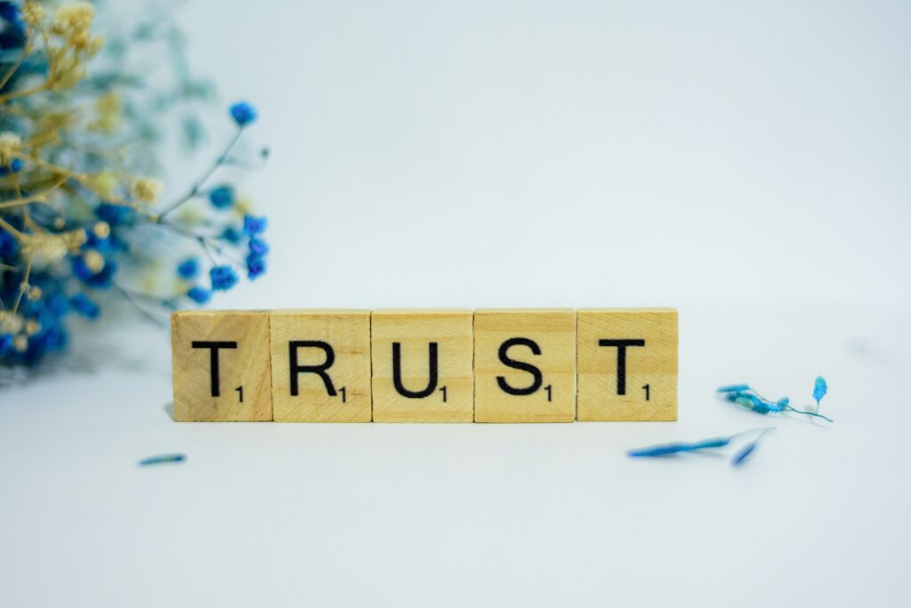 Wooden letter tiles spell 'TRUST' on a light surface with blue flowers blurred in the background, giving a soft, airy feel.