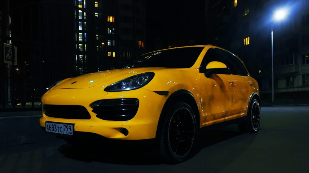 Front three-quarter view of a bright yellow luxury car parked on a dark city street at night, with lit windows and a streetlight in the background.
