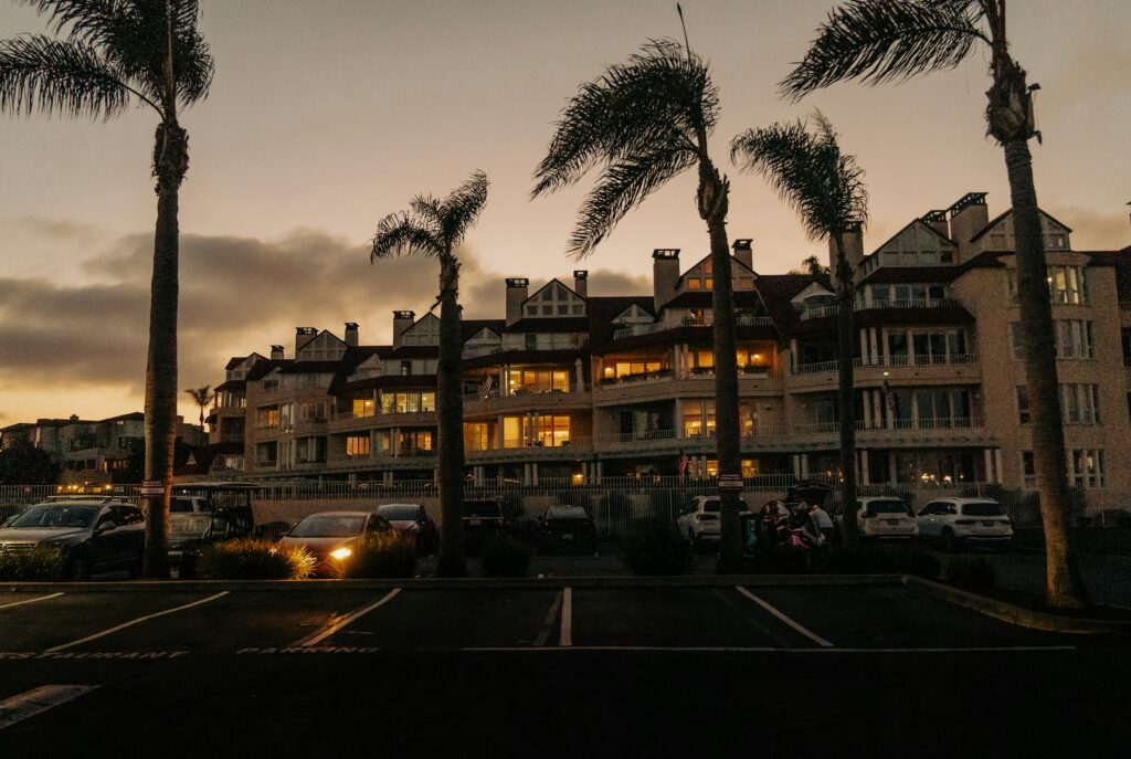 Sunset view of a row of beachside condo buildings with palm trees in the foreground and warmly lit balconies and windows.