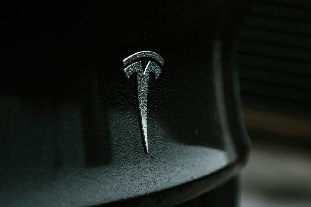Close-up of a rain-speckled Tesla logo on a dark car hood.