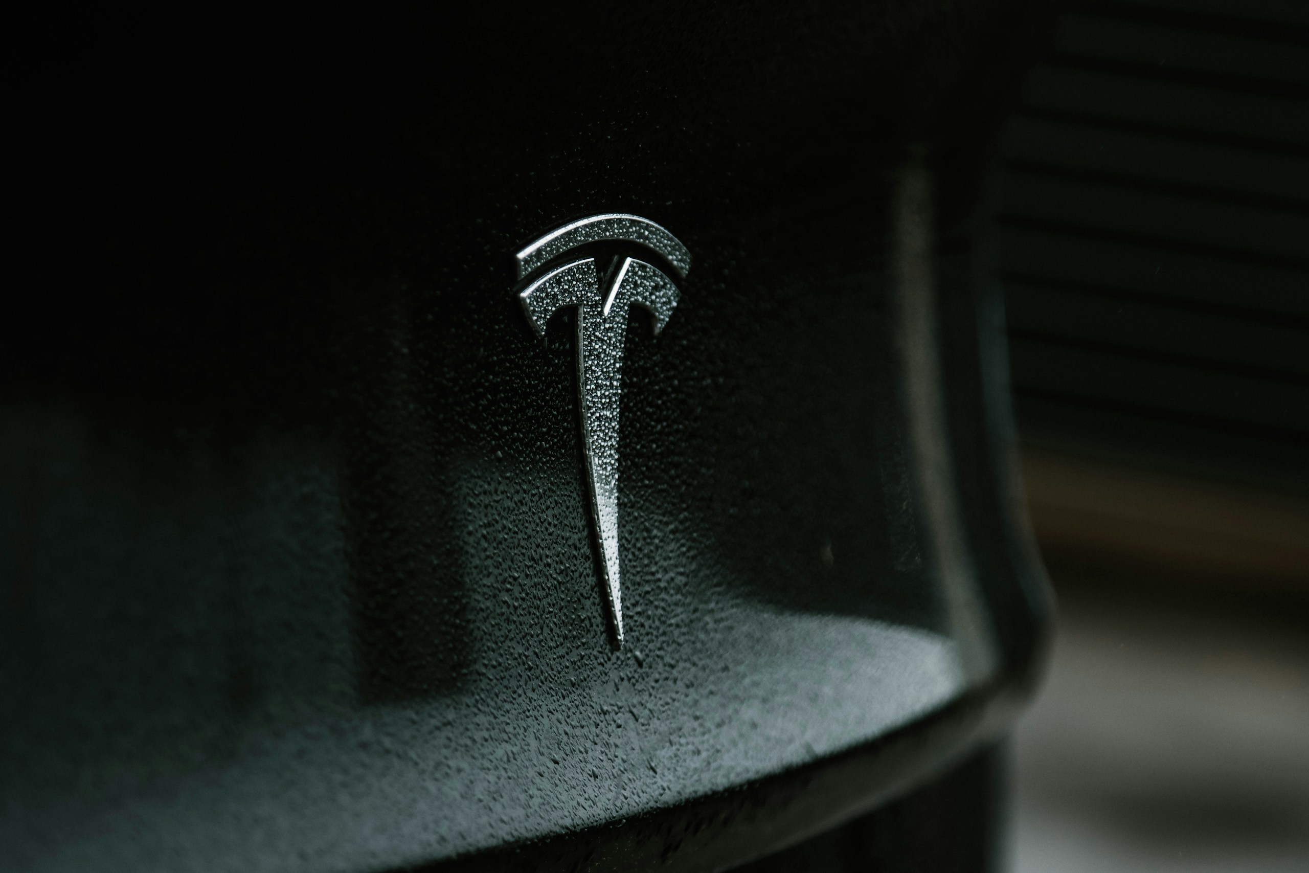 Close-up of a rain-speckled Tesla logo on a dark car hood.