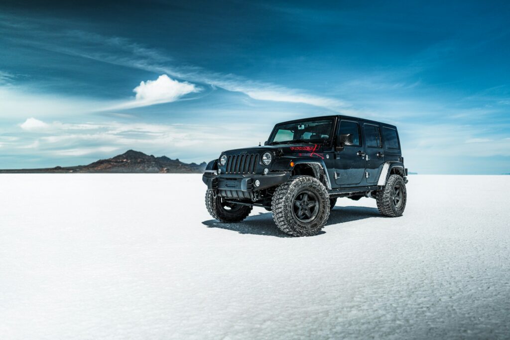 Black Jeep Wrangler parked on a snow-covered landscape with a blue sky and distant mountain range in the background.