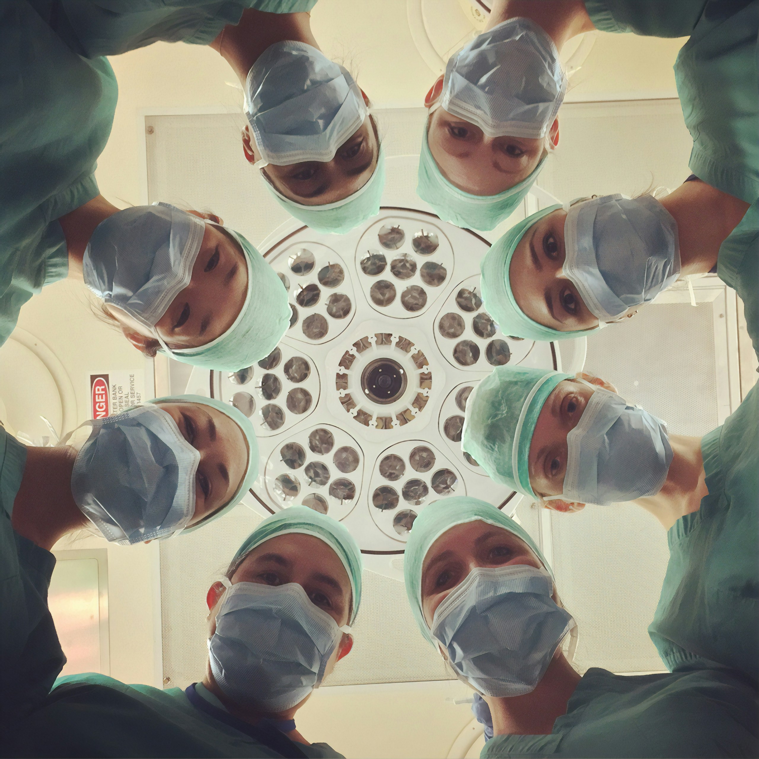 Surgeons in green scrubs and masks form a circle around the camera in an operating room overhead lights above them