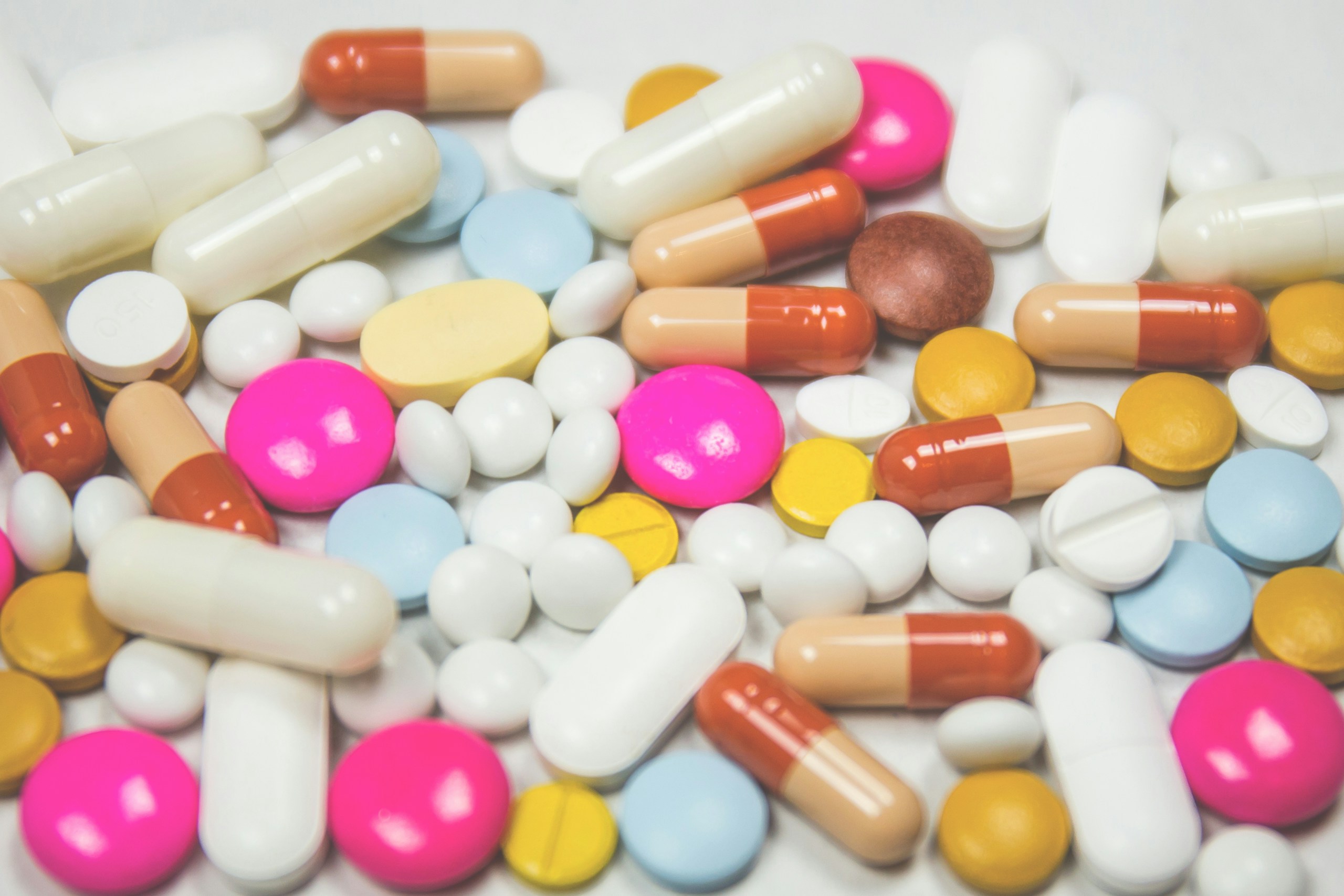 Close-up of a variety of colorful capsules and tablets scattered on a white surface, like pills and meds mixed together.