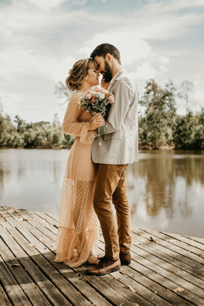 Couple embracing on a wooden dock by a calm lake, woman in a peach dress holding a bouquet, man in a light blazer.