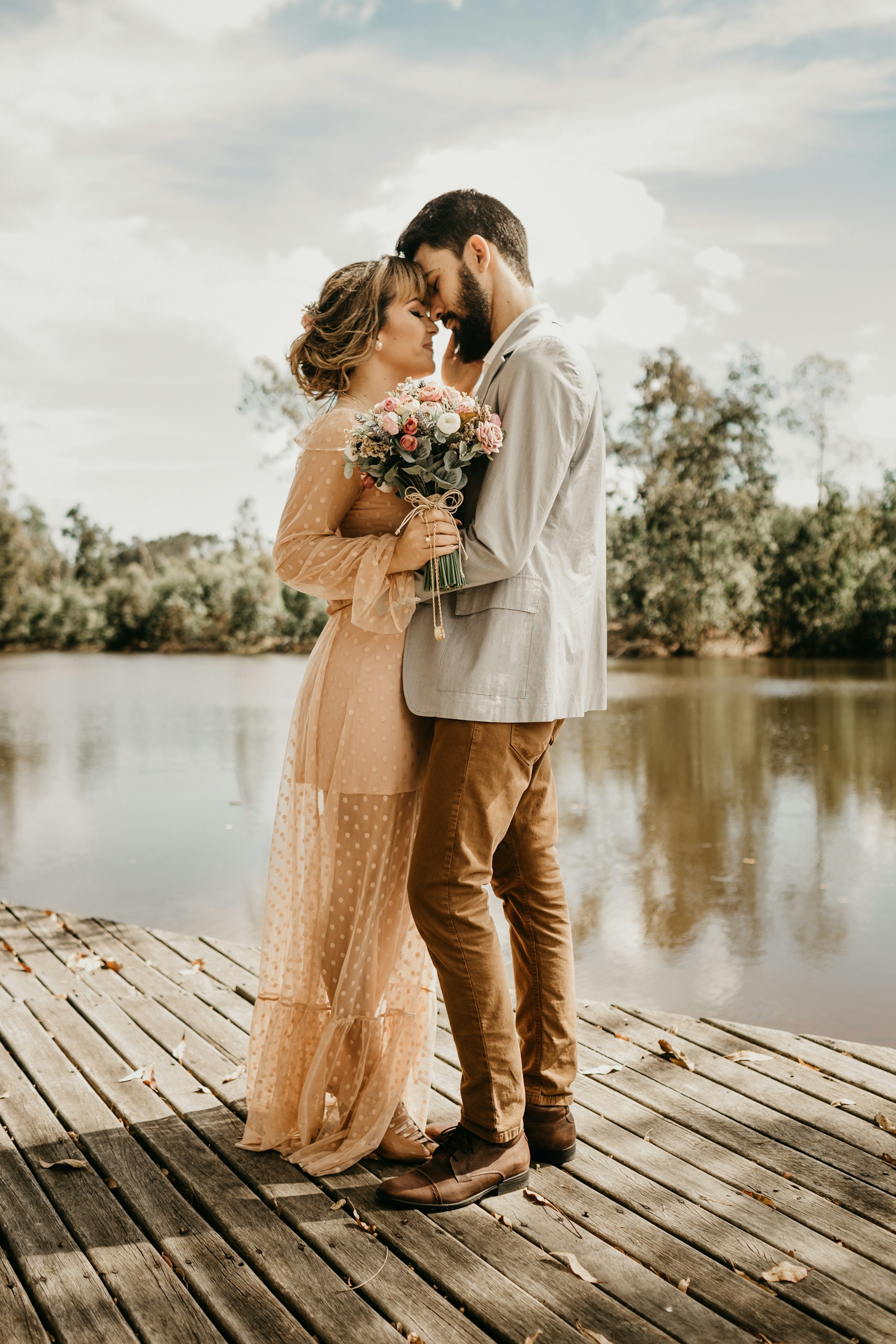 Couple embracing on a wooden dock by a calm lake, woman in a peach dress holding a bouquet, man in a light blazer.