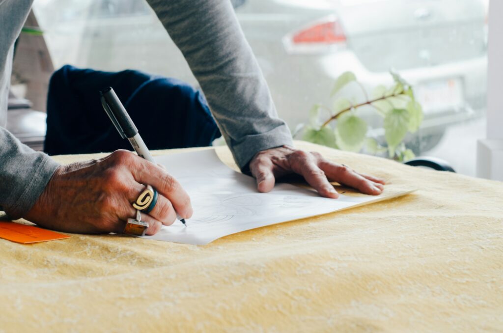 Close-up of hands sketching on white paper with a black marker on a yellow tabletop.