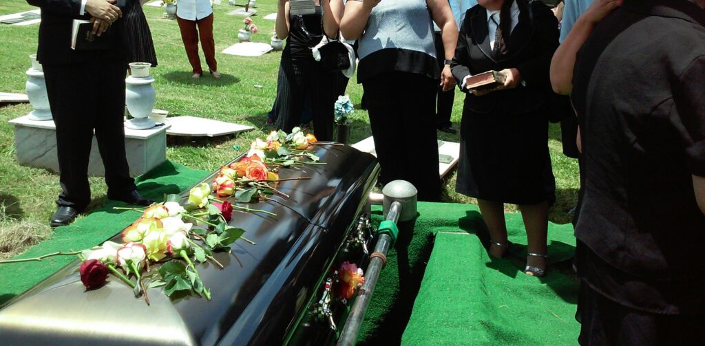 Casket decorated with colorful roses at a grave as mourners stand nearby.