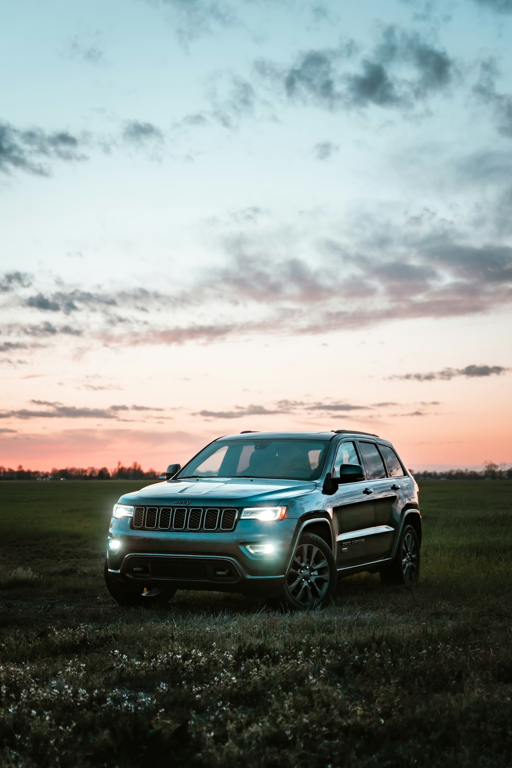 Silver Jeep SUV parked in a grassy field at sunset with headlights on.