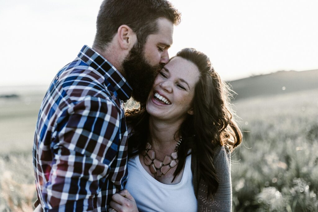 Man with a beard kissing the woman’s head as they embrace in a sunlit field; she smiles warmly outside.