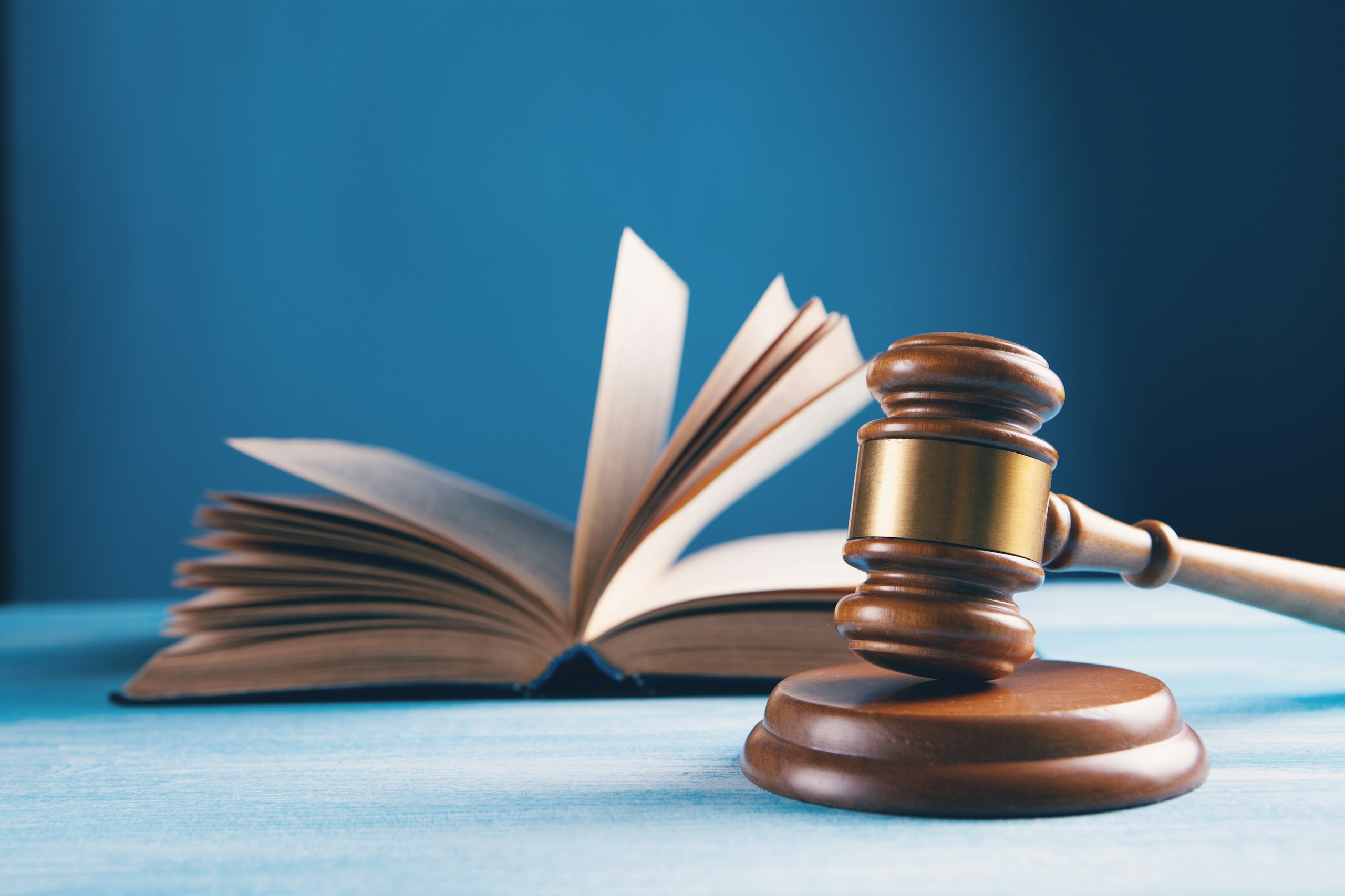 Judge's gavel resting on a wooden block with an open book in the background on a blue surface.