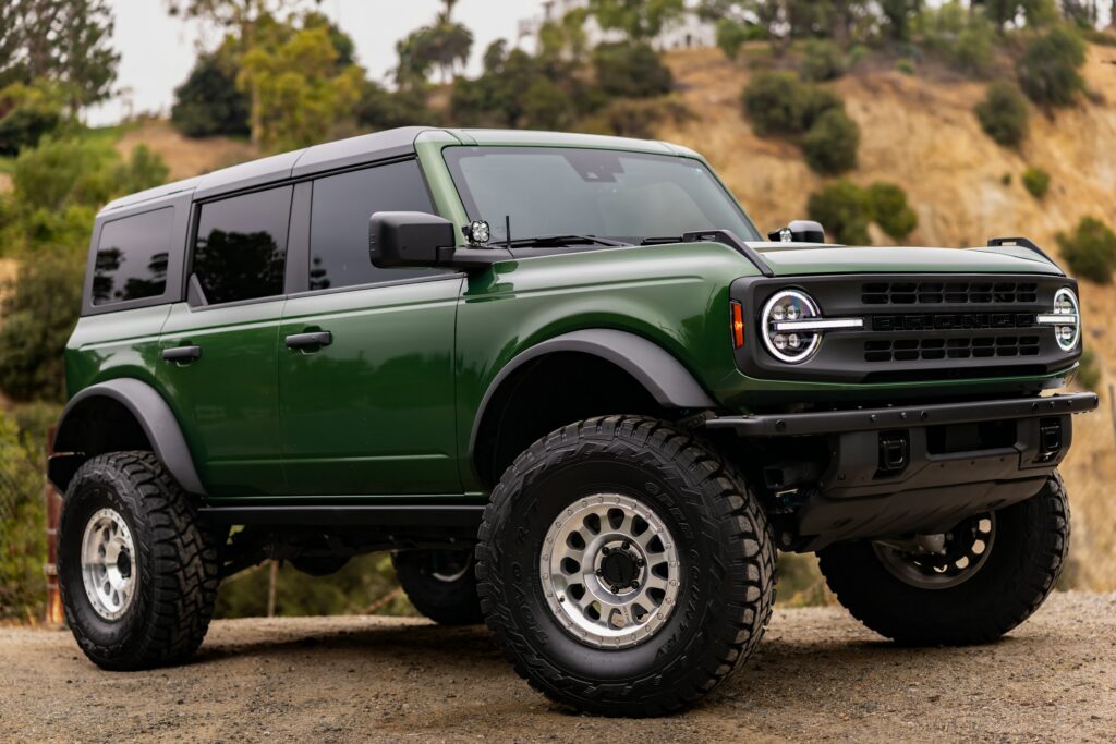 Green lifted off-road SUV with large mud-terrain tires on a dirt path, hillside backdrop.