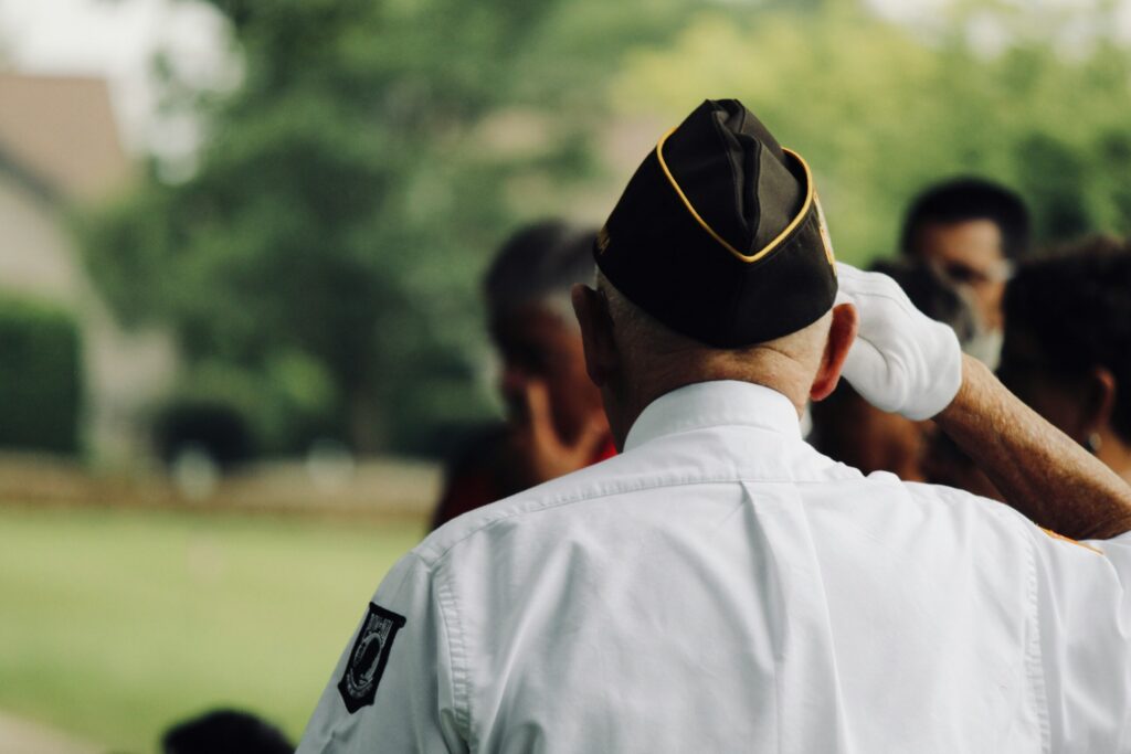 Back view of a person in a white uniform adjusting a black peaked cap outdoors. A few people and greenery are blurred in the background.