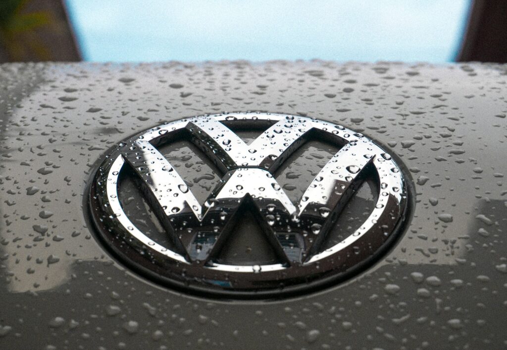 Close-up of a shiny car hood with water droplets and a chrome emblem in the center, likely a vehicle logo.