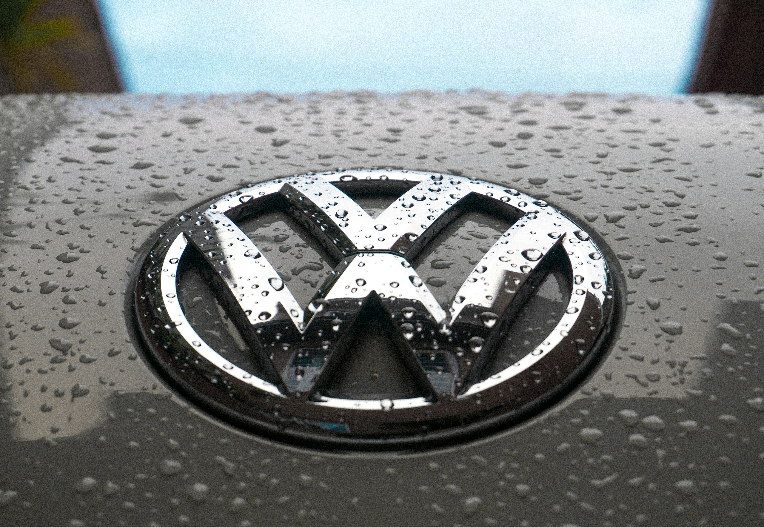 Close-up of a shiny car hood with water droplets and a chrome emblem in the center, likely a vehicle logo.