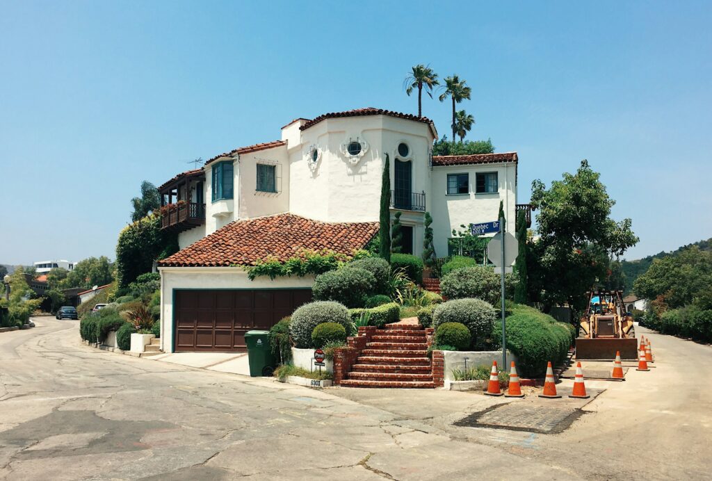 Corner view of a Spanish-style white house with red tile roof on a sunny street; construction cones line the right sidewalk with equipment in the distance.
