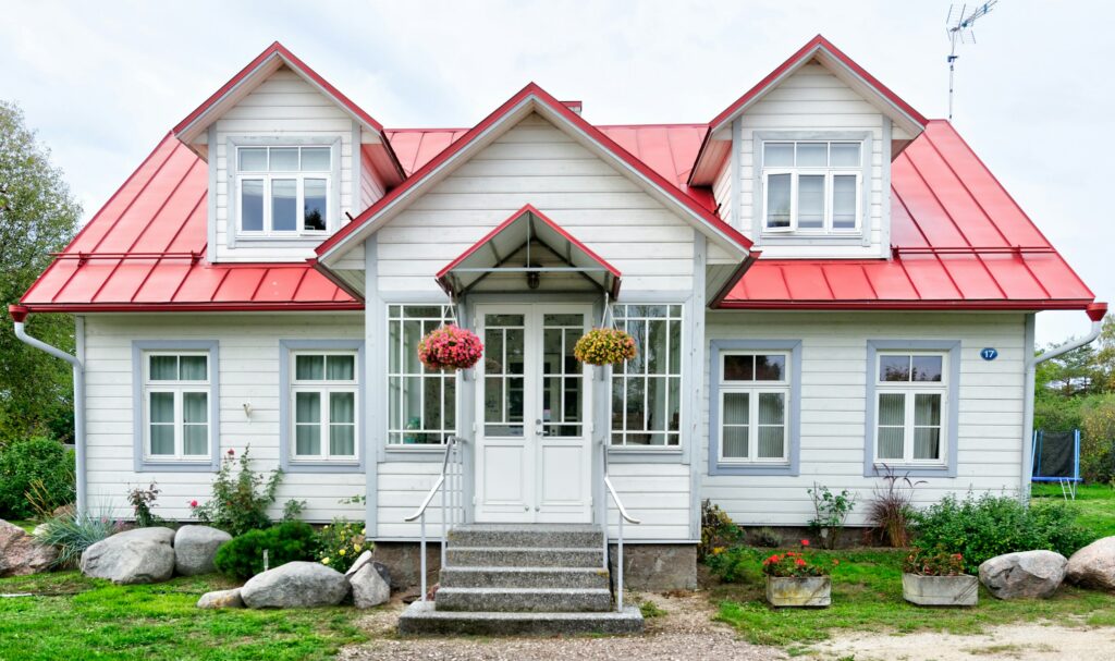 White wooden house with a bright red metal roof, central glass doors, and two hanging flower baskets along the entryway, surrounded by a garden and rocks.