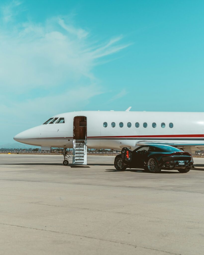 Airplane parked on tarmac with stairs extended to the door and a dark sports car beside it under a clear blue sky.