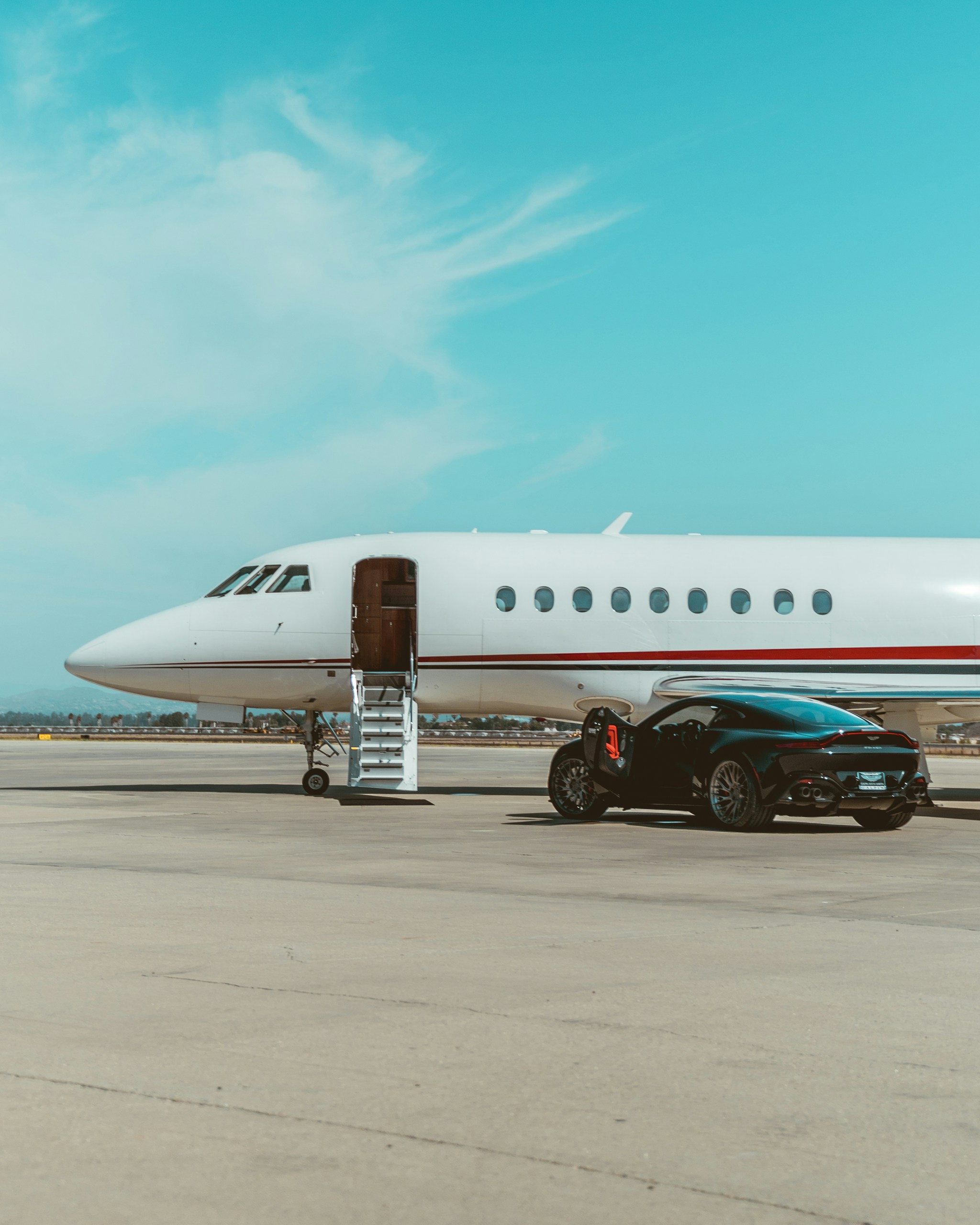 Airplane parked on tarmac with stairs extended to the door and a dark sports car beside it under a clear blue sky.