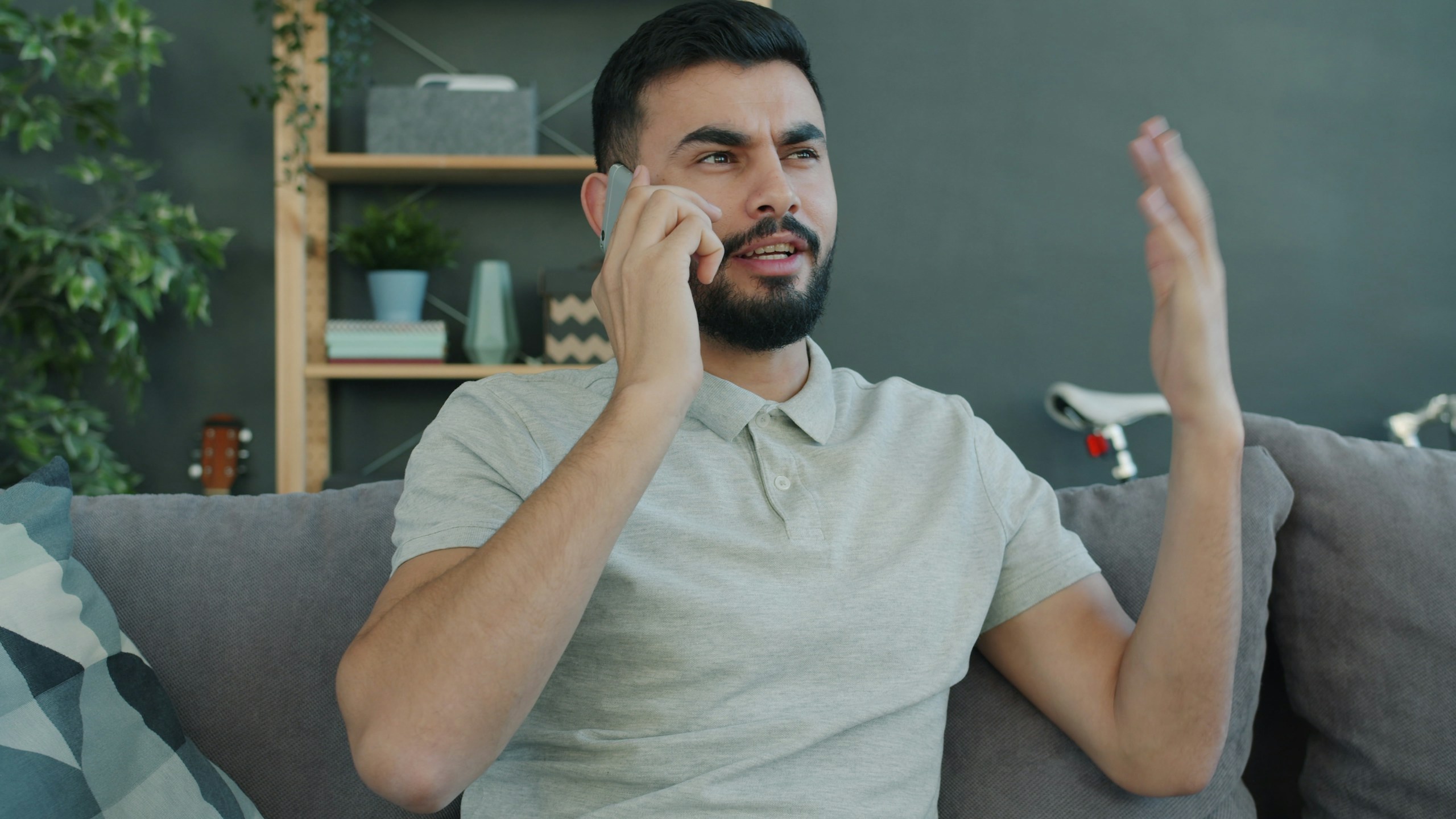 Joyful Arabian guy is speaking on mobile phone gesturing sharing emotions at home sitting on sofa in cozy studio apartment. People and lifestyle concept.